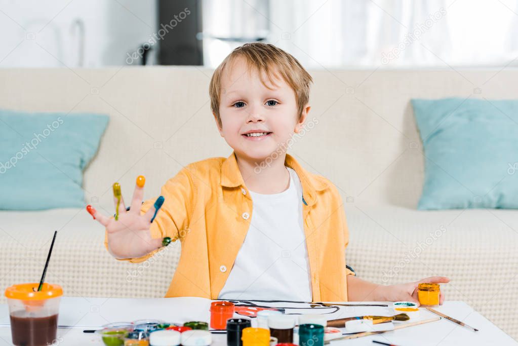 adorable preschooler boy looking at camera and showing painted palm during drawing at home