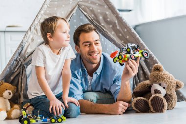 father and preschooler son playing with toy car under wigwam at home clipart