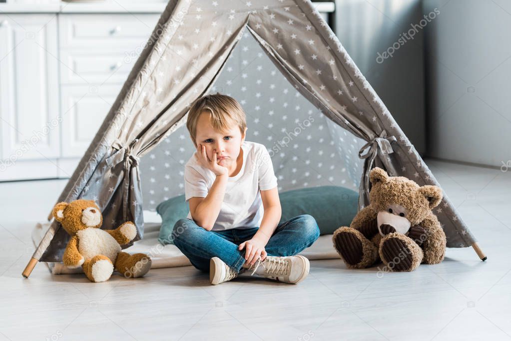 adorable sad preschooler boy sitting with teddy bears and propping chin with hand in wigwam at home