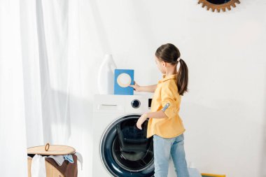 side view of child in yellow shirt near washer taking washing powder in laundry room clipart