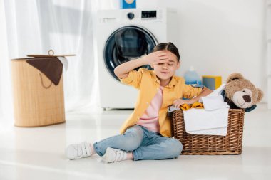 Sarı tişört ve kot pantolon ile çamaşır odasında ayı oyuncak sepeti oturan yorgun çocuk