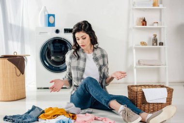 confused woman sitting on floor near scattered clothes and baskets in laundry room clipart