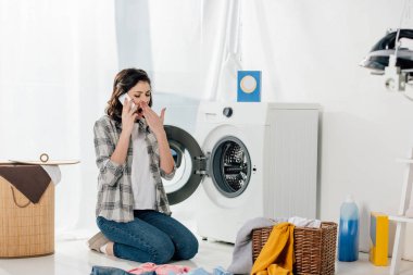 woman sitting near washer, talking on smartphone and yawning in laundry room clipart