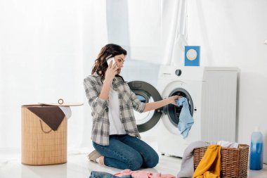 woman putting clothes in washer and talking on smartphone in laundry room clipart