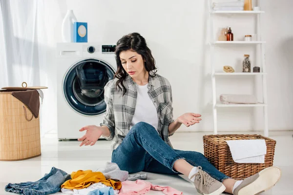 confused woman sitting on floor near scattered clothes and baskets in laundry room