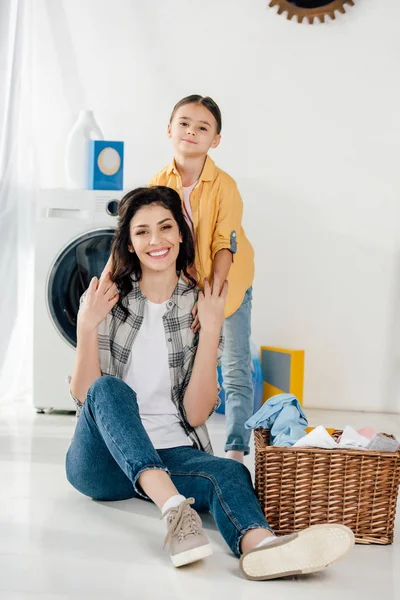 daughter in yellow shirt holding mother sitting on floor near basket in laundry room