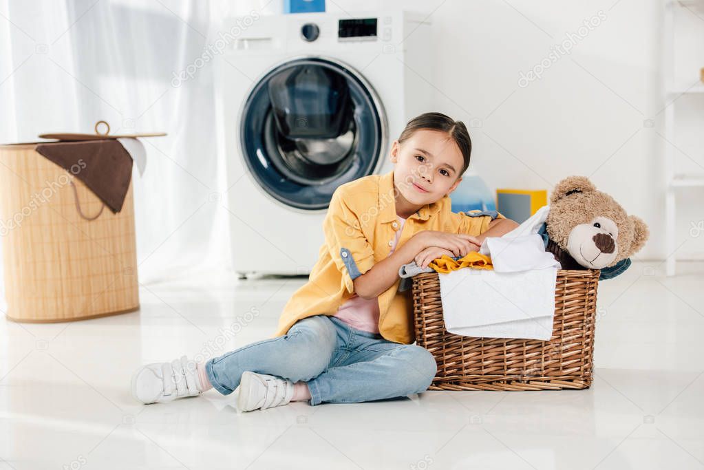 child in yellow shirt and jeans sitting near basket with bear toy in laundry room