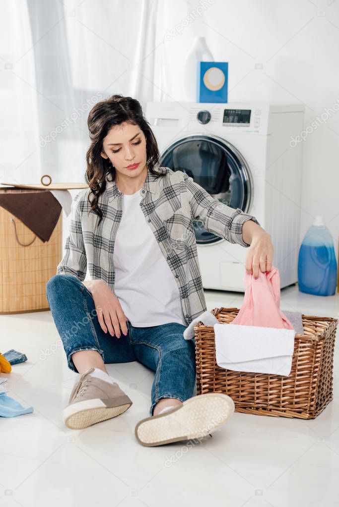 attractive woman sitting on floor and putting clothes to basket in laundry room