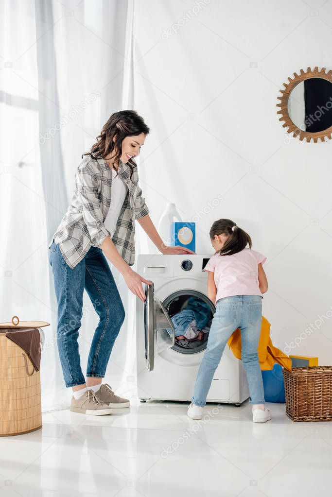 daughter in pink t-shirt putting clothes in washer wile mother standing near in laundry room