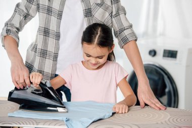 mother and daughter in pink t-shirt ironing in laundry room clipart