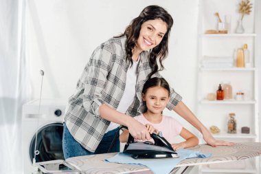 smiling mother in grey shirt and daughter in pink t-shirt ironing in laundry room clipart