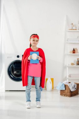 child standing in red homemade superhero suit and holding washing powder in laundry room clipart