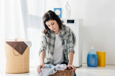 selective focus of upset woman in grey shirt sitting near basket in laundry room clipart