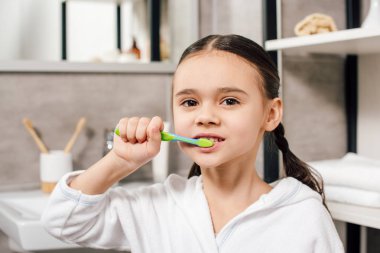 selective focus of child in white bathrobe brushing teeth in bathroom  clipart