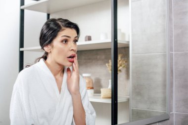 agitated woman in white bathrobe looking to mirror in bathroom clipart