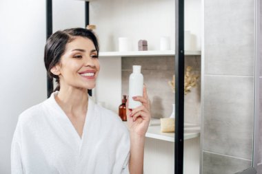 aynaya bakarak, losyon ile şişe tutan ve banyoda gülümseyen beyaz bornoz kadında