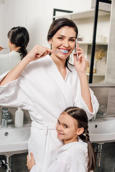 Filha Abraçando Mãe Escovando Dentes Falando Smartphone Banheiro — Fotografia de Stock