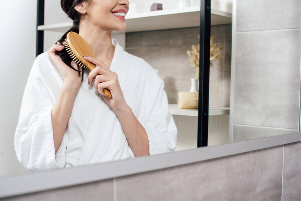 cropped view of woman in white bathrobe combing hair and looking to mirror in bathroom