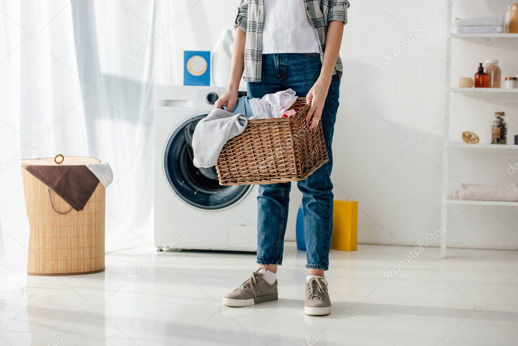 cropped view of woman in grey shirt and jeans holding basket in laundry room