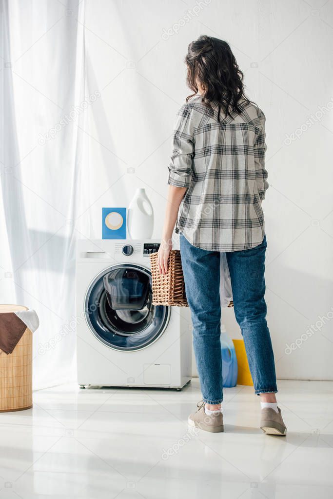 back view of woman in grey shirt and jeans holding basket near washer in laundry room