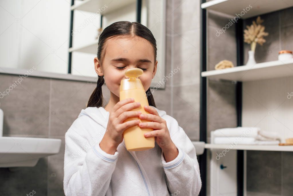 cute child in white bathrobe sniffing shower gel in bathroom 