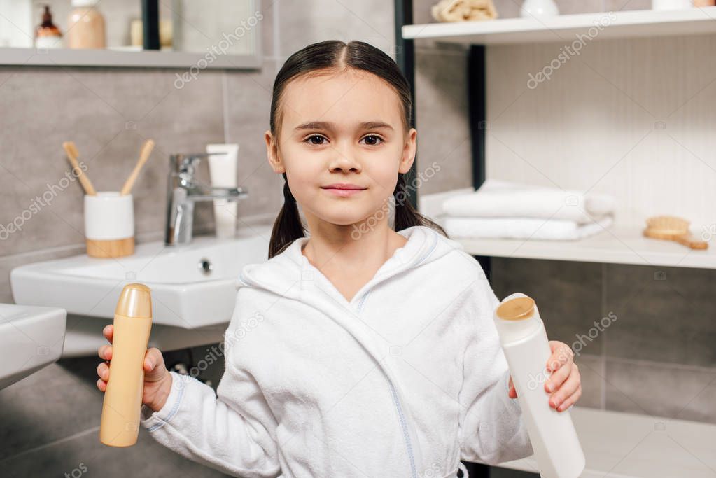 cute child in white bathrobe holding bottles with shower gel in bathroom 
