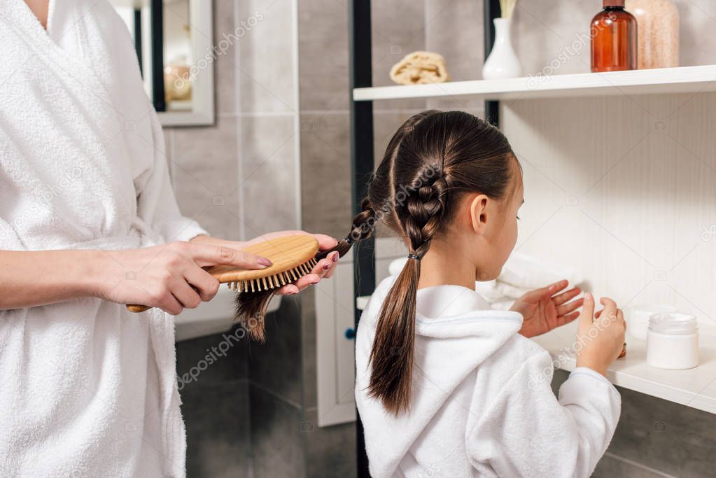 mother in white bathrobe combing daughter hair near shelves in bathroom