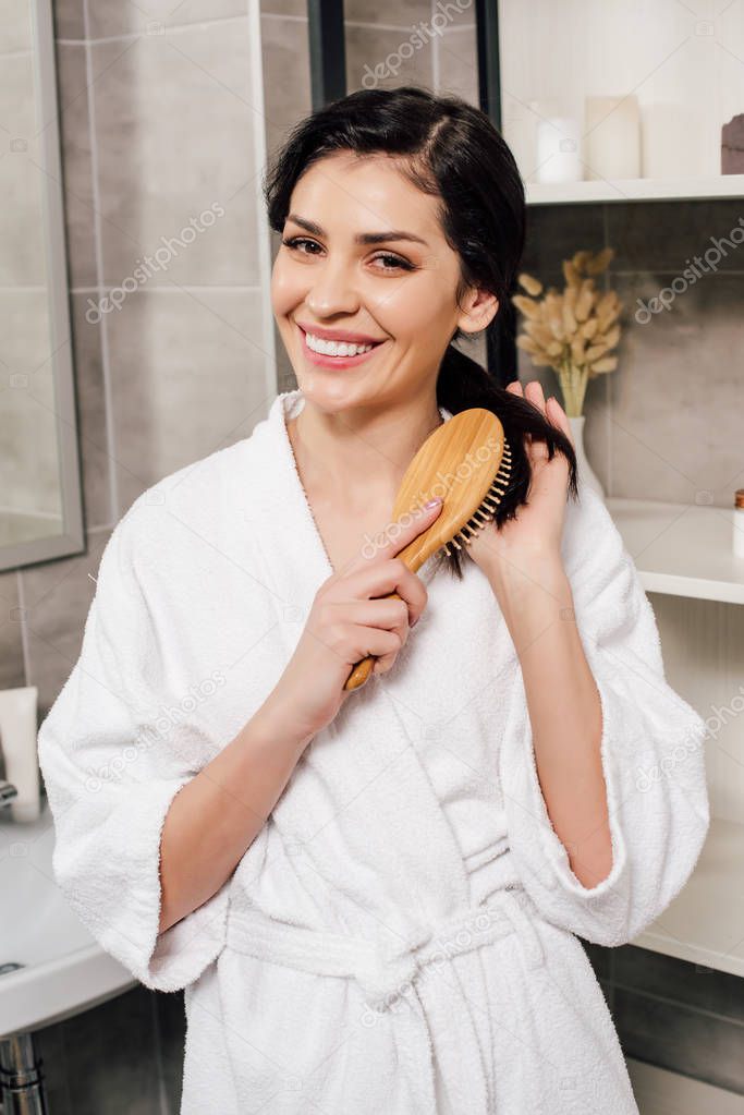 woman in white bathrobe combing hair and smiling in bathroom