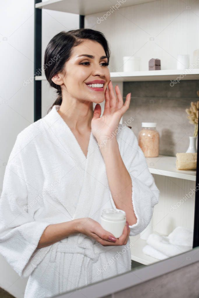 woman in white bathrobe looking to mirror and applying cosmetic cream in bathroom
