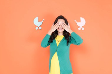 Glad pregnant woman in green cardigan covering eyes with hands on orange background clipart