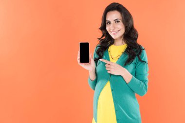 Boş ekran üzerinde turuncu izole ile smartphone, parmağınızla işaret memnun hamile kadın