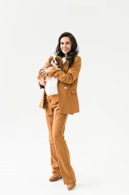 Kahverengi elbiseli köpek beyaz arka plan üzerinde tutarak gülümseyen hamile kadın