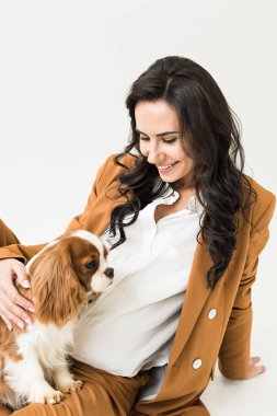 Laughing pregnant woman in brown jacket stroking dog on white background clipart