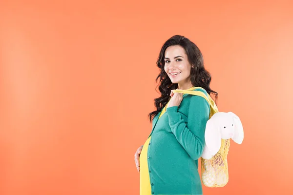 笑顔の妊婦グッズとひも付きの袋を押しながらオレンジに分離されて腹に触れる — ストック写真
