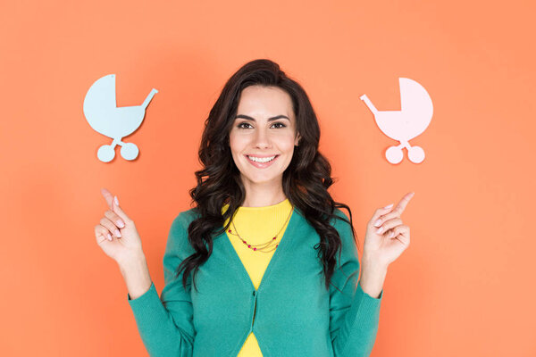 Blissful pregnant woman in green cardigan pointing with fingers at paper strollers on orange background