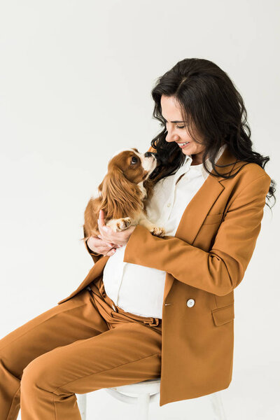 Laughing pregnant woman in brown suit looking at dog isolated on white 