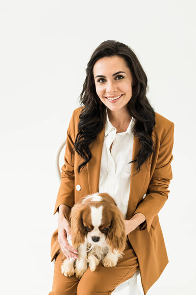 Glad brunette woman in brown suit holding dog and smiling isolated on white