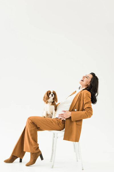 Blissful pregnant woman sitting on chair with dog and laughing on white background