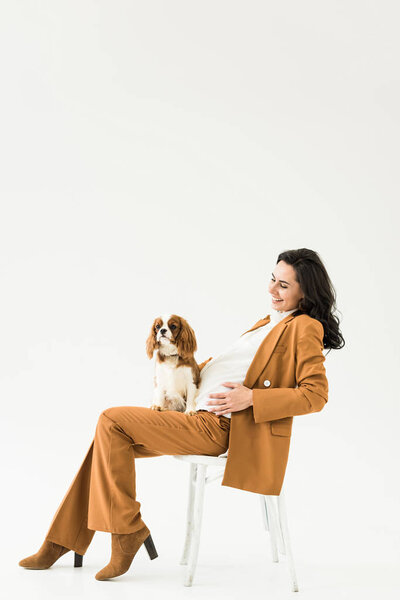 Joyful pregnant woman in brown suit sitting on chair with dog on white background