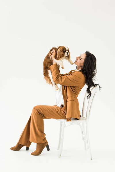Pregnant woman in brown suit sitting on chair and holding dog with smile on white background