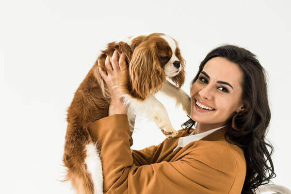 Cheerful brunette woman in brown jacket holding dog isolated on white