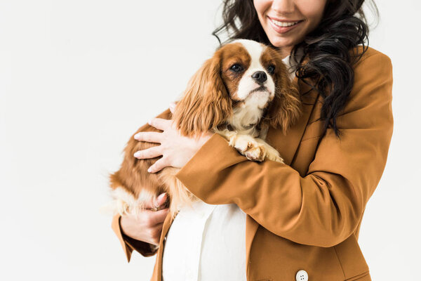 Cropped view of laughing pregnant woman in brown jacket holding dog isolated on white
