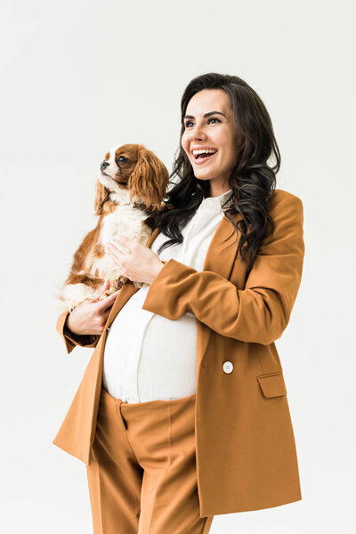 Blissful pregnant woman in suit holding dog isolated on white