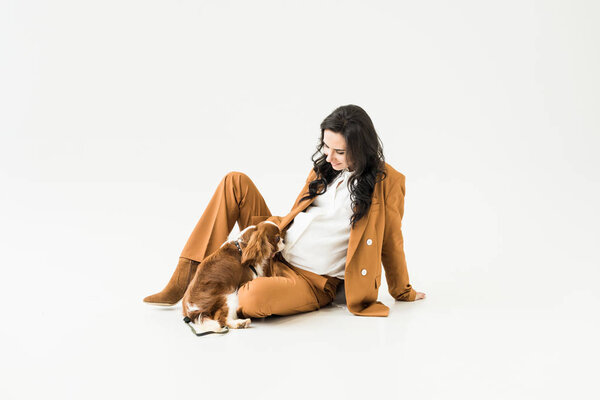 Stylish pregnant woman in suit sitting on floor and looking at dog on white background