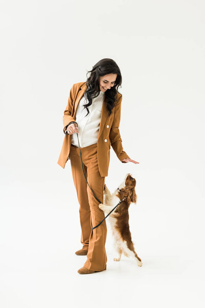Smiling pregnant woman in brown suit playing with dog on white background