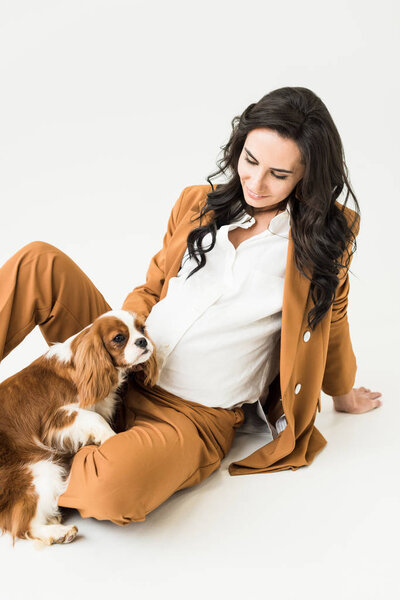 Charming pregnant woman sitting on floor and looking at dog on white background