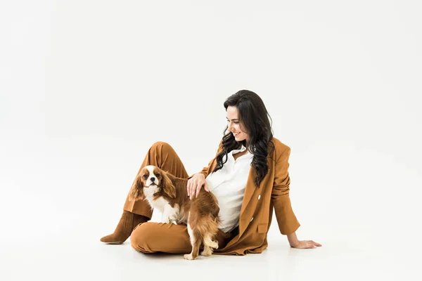 Elegant pregnant woman sitting on floor and stroking dog on white background