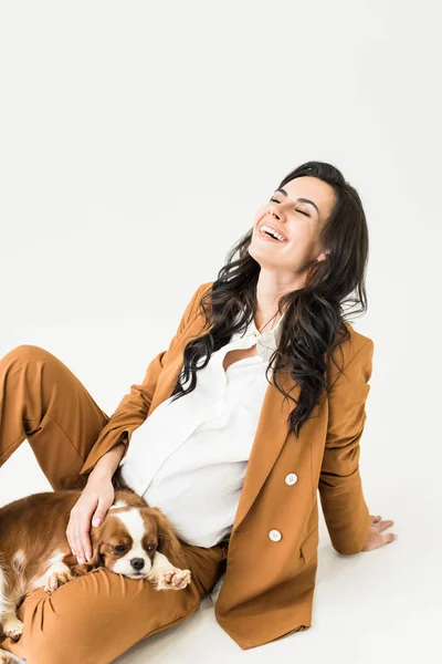 Pregnant woman stroking dog and laughing with closed eyes on white background