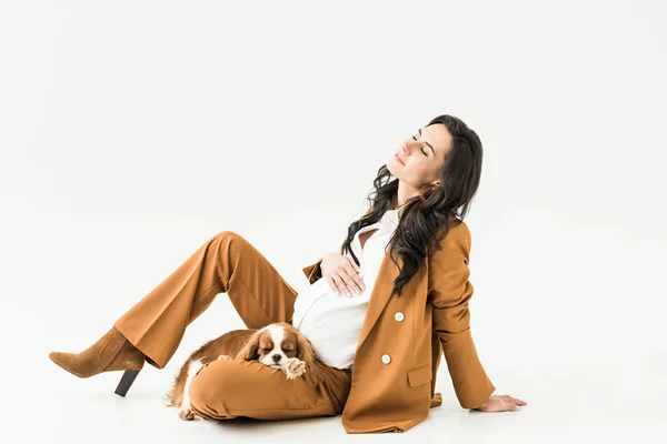 Dreamy pregnant woman sitting on floor with sleeping dog on white background