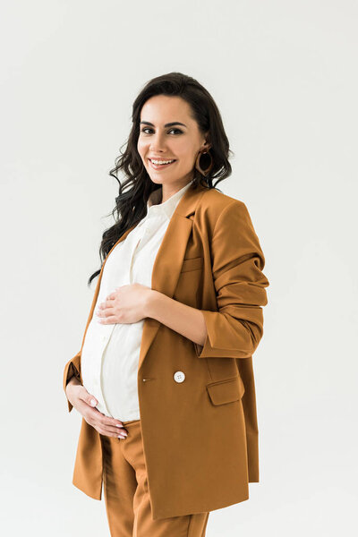 Blissful pregnant woman in brown suit touching tummy and looking at camera isolated on white